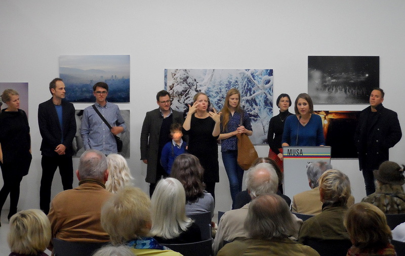 Gunda Achleitner, die Kuratorin der aktuellen Ausstellung im musa bei der Eröffnungsrede, im Hintergrund einige der teilnehmenden KünstlerInnen, an der Wand Arbeiten von Lina Scheynius, Sarajevo (die Künstlerin in Jeans mit brauner Tasche)